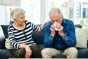 senior-woman-comforts-husband-on-couch-as-he-blows-his-nose