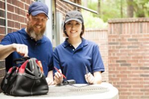 man-and-woman-technicians-wearing-blue-shirts-and-caps-smiling-and-fixing-outside-ac-with-toolkit